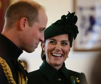 Two people smiling, dressed in formal attire, one wearing a green hat with a floral accessory.