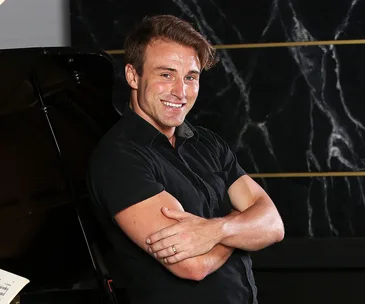 Man in a black shirt smiling, standing with arms crossed in front of a dark, marble-patterned background.