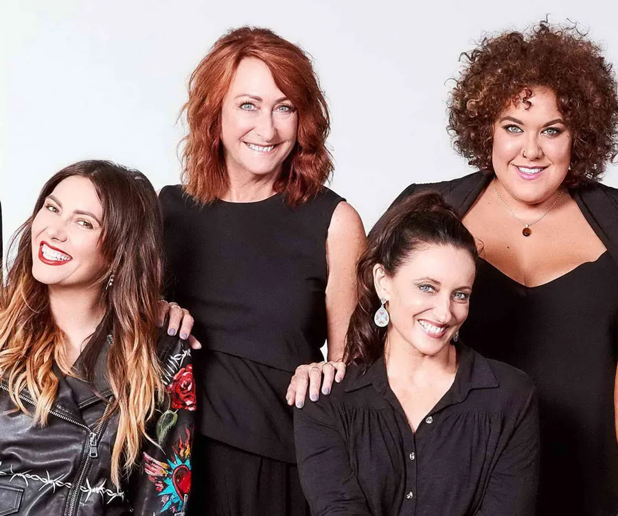 Four women smiling together, all wearing black outfits against a plain backdrop.