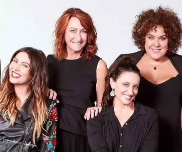 Four women smiling together, all wearing black outfits against a plain backdrop.
