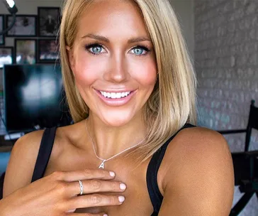 Blonde woman with blue eyes smiling, wearing a black top and "A" necklace, indoors with a blurred background.