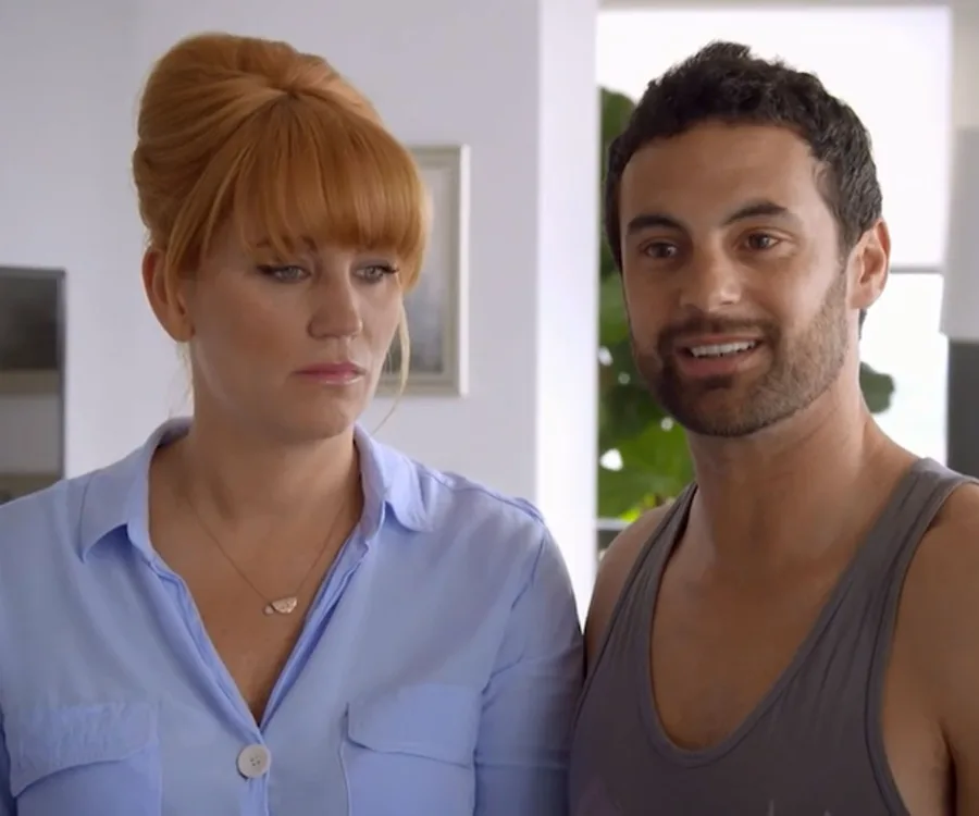 Two people standing together indoors; the woman looks serious while the man is smiling.
