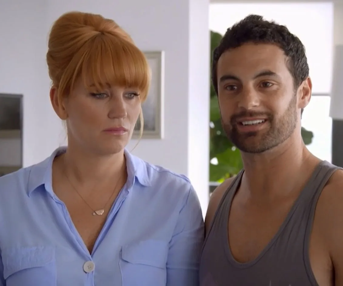 Two people standing together indoors; the woman looks serious while the man is smiling.