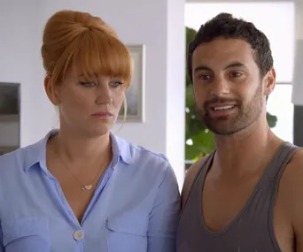 Two people standing together indoors; the woman looks serious while the man is smiling.