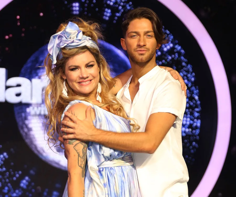 Couple in elegant attire poses on stage set of "Dancing with the Stars," surrounded by glittering lights.