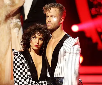 Dancers embrace on stage in themed outfits, one in black and white checkered design, with dramatic red lighting behind.