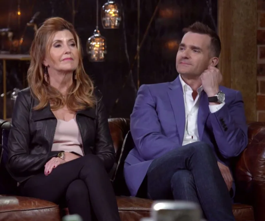 Two people sitting on a sofa, one wearing a black jacket, the other in a blue blazer, in a warmly lit room.