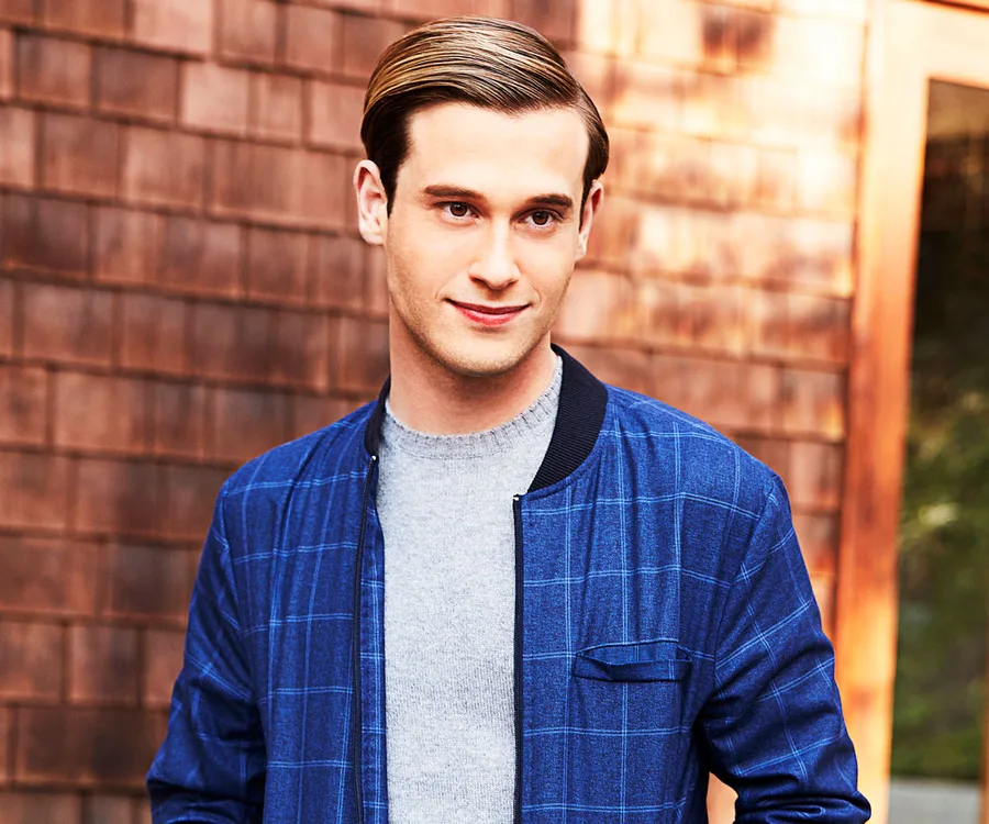 Young man in a blue plaid jacket standing outdoors against a wooden wall.