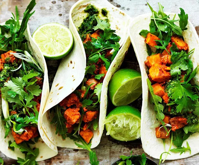 Three sweet potato tacos with greens and lime wedges on a metal tray.