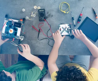 Two children working on robotics projects with tools and a tablet on a table, viewed from above.