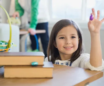 A smiling girl holds up a purple egg during an indoor Easter egg hunt, with a basket and books nearby.