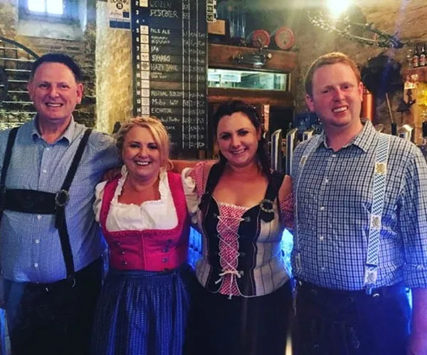 Group of four people in traditional Bavarian outfits, smiling inside a bar with a chalkboard menu behind them.