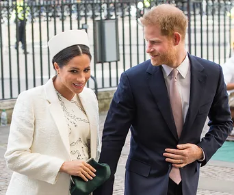 A man in a suit and a woman in a white coat and hat, smiling, walking together outdoors.