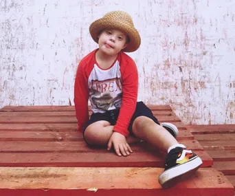 Young child with Down syndrome wearing a straw hat and red shirt sits on a wooden platform, smiling gently.