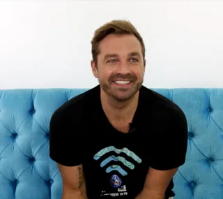 Man with brown hair and beard, wearing a black T-shirt, smiling while sitting on a blue couch.