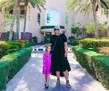 Woman and child in colorful clothes stand on path in front of a pink building with palm trees and manicured hedges.