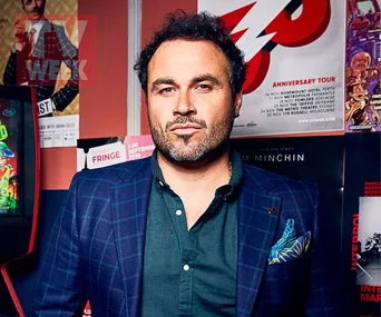 Miguel Maestre in a blue plaid suit jacket and green shirt, standing against a wall with event posters.