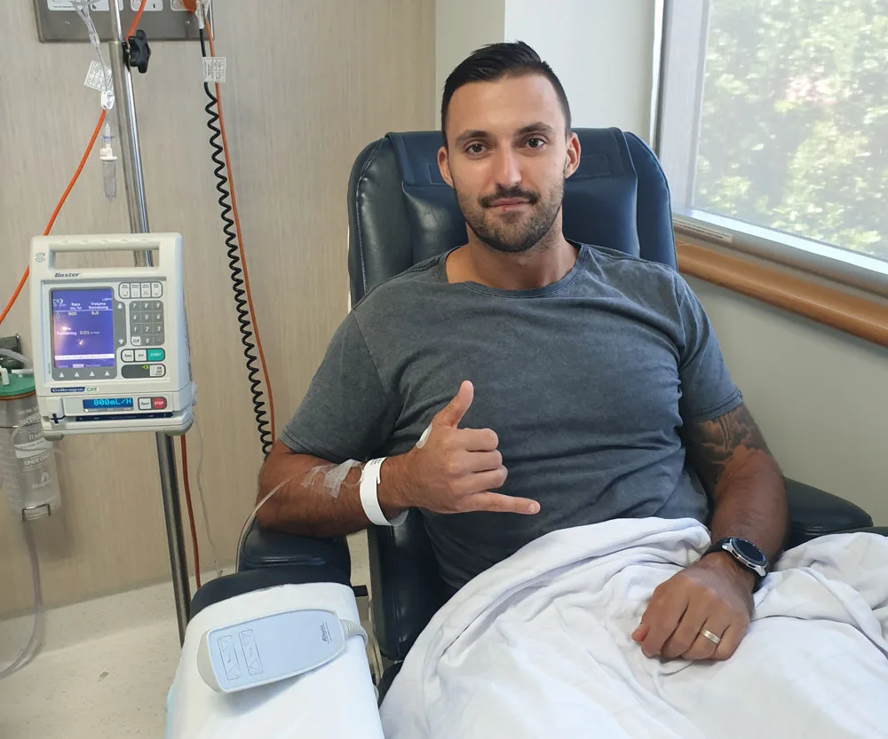 Man in a hospital setting giving a shaka sign while seated, with IV drip beside him.