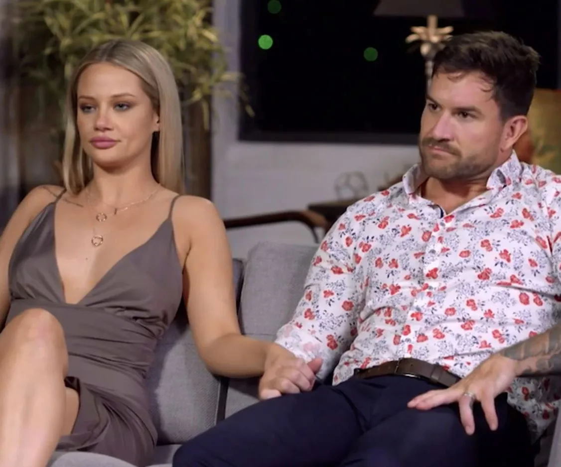 A couple sitting on a couch, holding hands, with neutral expressions; indoor setting.