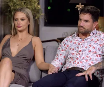 A couple sitting on a couch, holding hands, with neutral expressions; indoor setting.