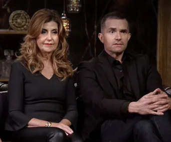 Two people sitting together on a couch, wearing dark clothing, looking serious.