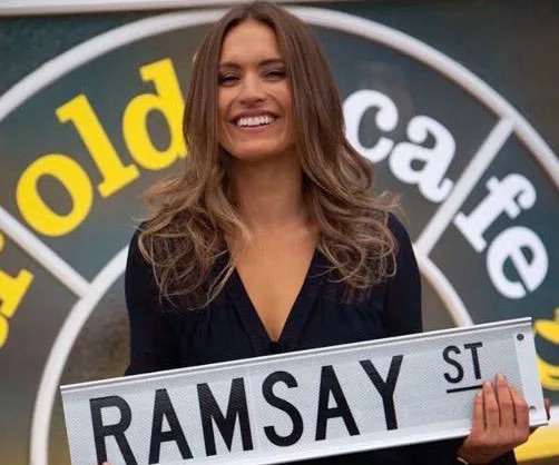 A woman smiles holding a "Ramsay St" sign, with a blurred cafe logo behind her, related to "Neighbours" series.