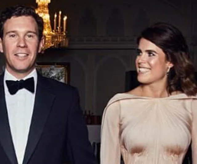 A couple in formal attire, with the woman in a Zac Posen dress, standing in an elegant room with a chandelier.