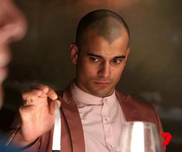 Man in a pink suit holds a knife intently at a dining table, with blurred figure in foreground. Channel 7 logo in corner.