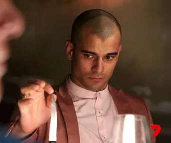 Man in a pink suit holds a knife intently at a dining table, with blurred figure in foreground. Channel 7 logo in corner.