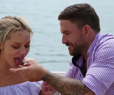 Couple on a date by the water, man feeds woman a snack, both smiling.