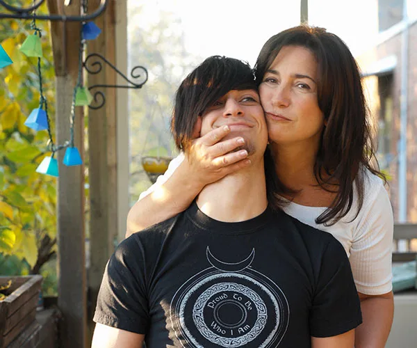 A woman playfully holds a young man's face on a porch, both smiling, surrounded by soft daylight.