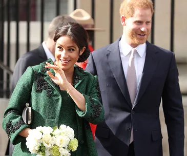 Two people smiling, with one person waving and holding a bouquet. They are dressed in formal attire outdoors.