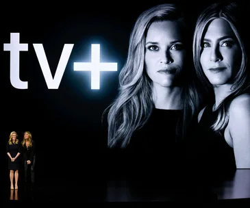 Reese Witherspoon and Jennifer Aniston on stage at Apple TV+ event, with their large photo and TV+ logo in the background.