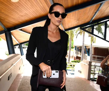 Woman in black outfit and sunglasses holding a phone and clutch, outside a building with a wooden roof.