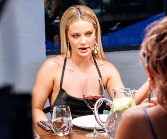 Woman with blonde hair in black top, dining with others, wine glass and a pitcher on table.