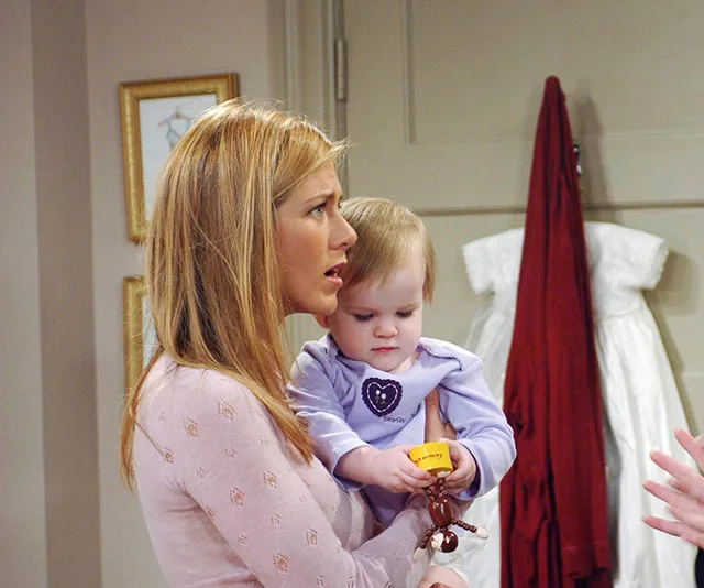 Rachel from "Friends" holds a baby wearing a heart sweater in a room with a red robe and white dress hanging on the wall.