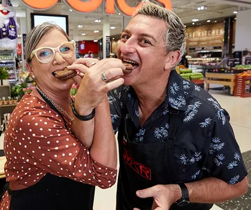 Two people in a supermarket wearing aprons, smiling and eating biscuits together.