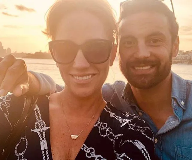 A couple taking a selfie at sunset near the water, with a city skyline in the background.