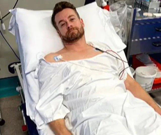 Man in a hospital bed wearing a gown, connected to medical monitors, looking towards the camera.