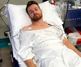 Man in a hospital bed wearing a gown, connected to medical monitors, looking towards the camera.