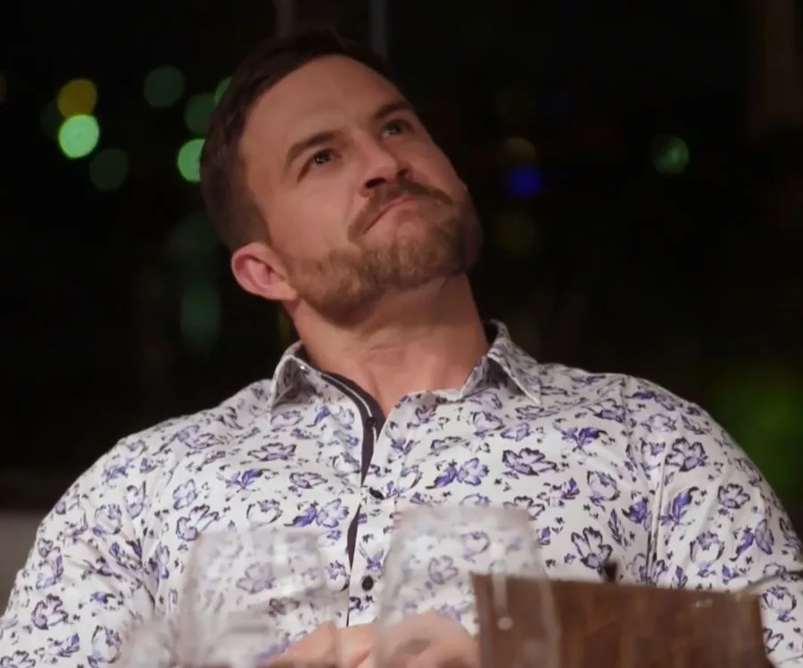 A man in a floral shirt gazes upwards reflectively in a dimly lit setting.