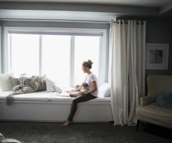 Woman sitting on a window seat with a laptop, holding a baby doll, white curtains in the background.