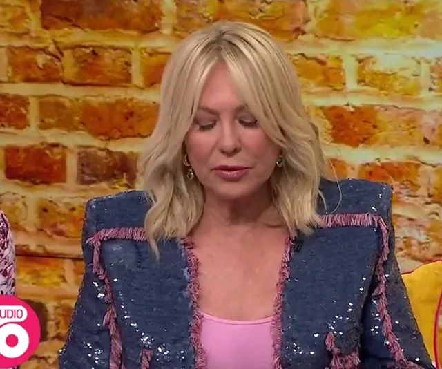A woman with blonde hair and a denim jacket, seated against a brick wall background on a talk show set.