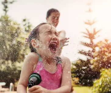 Child in pink swimsuit joyfully playing with water hose outdoors on a sunny day.