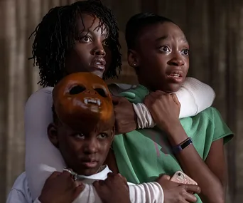 A woman and two children huddle closely, one wears a mask, all appear tense and concerned.