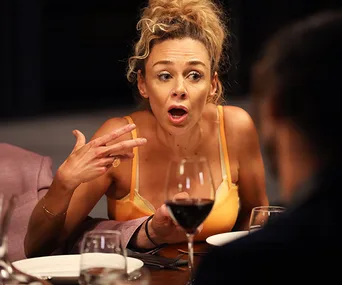 Woman with curly hair, in yellow top, animatedly gestures at dinner party.