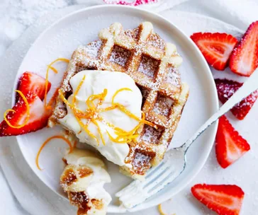 Waffle topped with whipped cream and orange zest, surrounded by sliced strawberries on a white plate.