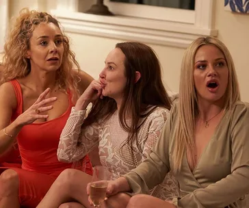 Three women sitting on a couch, looking surprised and engaged in a conversation.