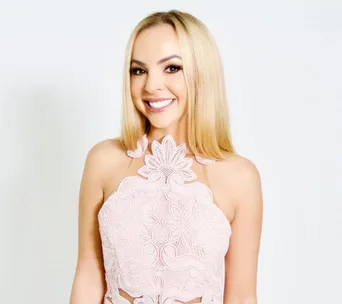 Smiling woman in a floral lace pink dress against a plain background.