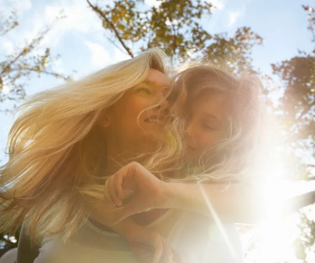 Mother with child on her back, smiling in sunlight, surrounded by trees and blue sky.